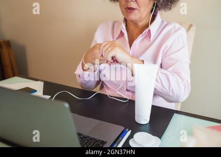 Crop content anziana imprenditrice femminile in auricolari applicazione crema idratante a portata di mano mentre si ha videochiamata sul portatile in ufficio Foto Stock