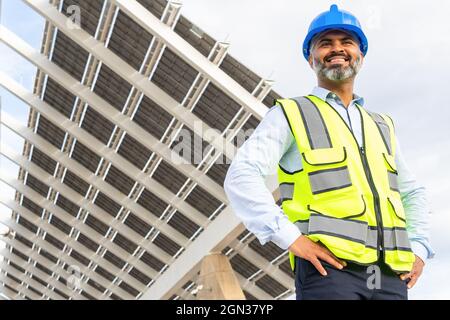 Da sotto di uomo etnia matura sorridente in hardhat e gilet in piedi vicino alla centrale solare Foto Stock