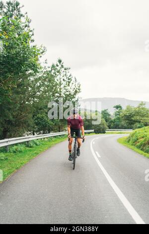 Sportivo in casco protettivo in bicicletta durante l'allenamento su strada asfaltata contro la collina verde e gli alberi sotto il cielo chiaro Foto Stock