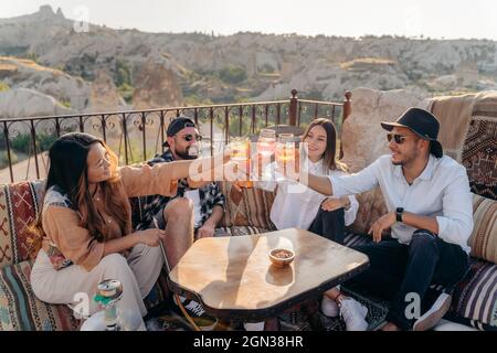 Un angolo alto di amici sorridenti seduti intorno al tavolo e alzando bicchieri di cocktail mentre sono appesi al bar in terrazza in Cappadocia, Turchia Foto Stock