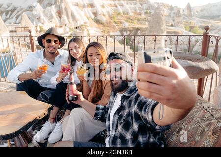 Amici maschili e femminili con cocktail e birra seduti sulla terrazza del bar e autoritratto su Instant photo camera in terrazza in Cappadocia, T. Foto Stock