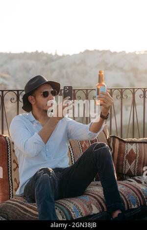 Uomo moderno in elegante vestito e occhiali da sole seduti su cuscini e fotografare una bottiglia di birra di vetro sul cellulare sul bar terrazza in Cappadocia, Foto Stock