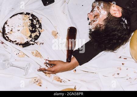 Da sopra di maschio ubriaco che dorme vicino a schiacciato torta di compleanno e bottiglia vuota durante la festa Foto Stock