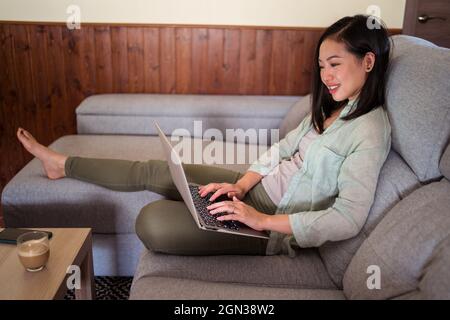 Vista laterale di una giovane e sorridente dipendente in remoto di etnia femminile che lavora su netbook sul divano contro il caffè e lo smartphone a casa Foto Stock