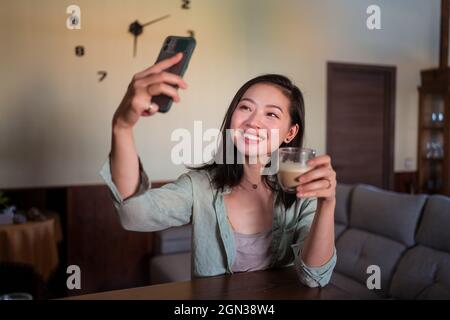 Contenuto giovane etnia femmina che prende autoritratto con caffè sul cellulare mentre si siede sul divano con il portatile a casa Foto Stock