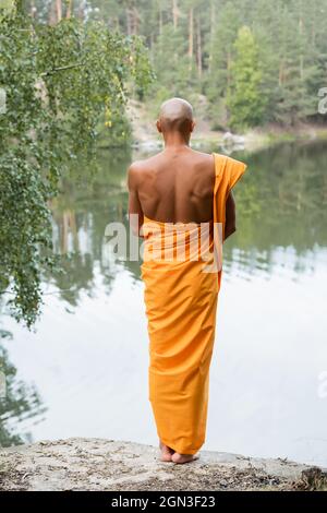 vista posteriore di uomo calvo in tradizionale accappatoio buddista meditando vicino stagno foresta Foto Stock