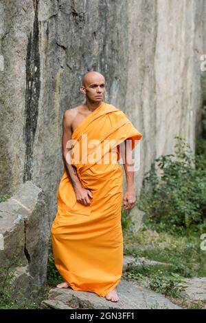 uomo in abiti buddisti tradizionali guardando lontano mentre in piedi vicino alla roccia Foto Stock