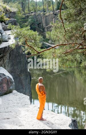 vista ad angolo alto del buddista in kasaya arancione meditando sulla scogliera rocciosa sopra l'acqua Foto Stock