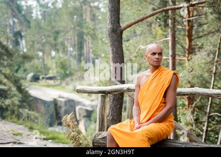 monaco buddista in tradizionale accappatoio arancione seduto su panca di legno nella foresta Foto Stock