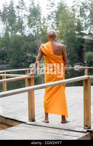 vista posteriore di uomo a piedi nudi in accappatoio buddista meditando su piattaforma di legno vicino al lago Foto Stock