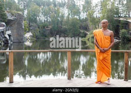 uomo in tradizionale accappatoio buddista in piedi su piattaforma di legno vicino al lago della foresta Foto Stock