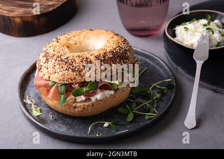 Bagel con formaggio cremoso, prosciutto secco e microgreens su un piatto su sfondo grigio, sandwich per la colazione, primo piano. Foto Stock