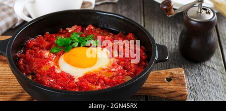 Shakshuka con salsa di pomodoro e uova fritte in una padella di ghisa per la colazione sul vecchio sfondo di legno. Stile rustico, fuoco selettivo. Foto Stock