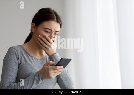 Sconvolto la donna graziosa millenaria depressa che copre la sua bocca con la mano e guardando il telefono Foto Stock