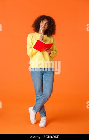 Full-length colpo verticale sognante e romantica ragazza carina rendendo la pianificazione, prendere appunti o elenco di cose da fare, imaging qualcosa come la scrittura in rosso carino notebook Foto Stock