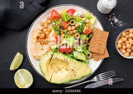 Insalata sana su piatto con avocado, hummus e verdure su un piatto, vista dall'alto. Sfondo di pietra nera Foto Stock