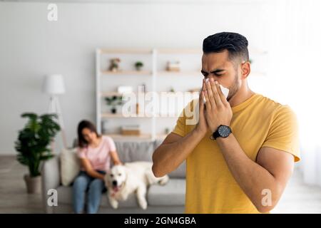 Il giovane ragazzo arabo starnutisce e soffia il naso in carta tissue, ha un cattivo freddo, rimanendo a casa con la sua ragazza e il suo cane Foto Stock