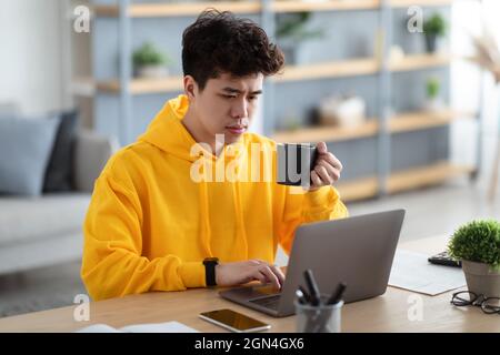 Uomo asiatico serio guardare video su pc, bere caffè caldo Foto Stock