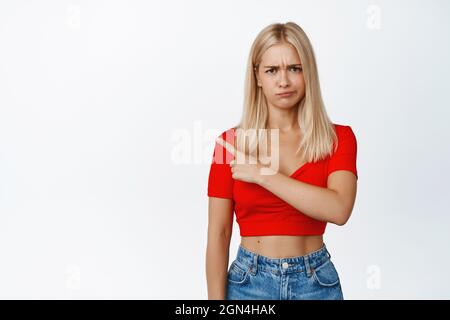 Donna arrabbiata accigliata puntando il dito a sinistra e accigliando, sconvolto su qualcosa, lamentandosi, in piedi su sfondo bianco Foto Stock