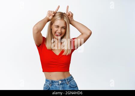 La ragazza bionda sassy mostra corna toro gesto e sorridente felice, in posizione spensierata su sfondo bianco Foto Stock