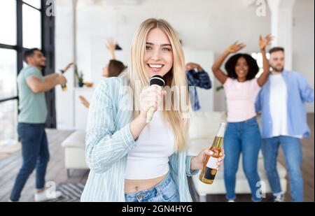 Donna bionda millenaria spensierata con una bottiglia di birra che canta il karaoke in festa con i suoi amici interrazziali a casa Foto Stock