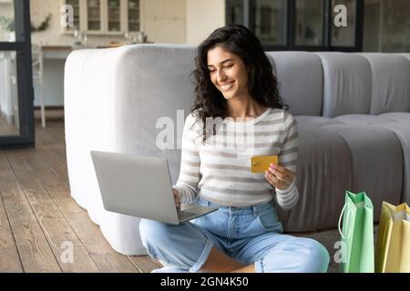 Cybershopping. Giovane signora caucasica con computer portatile, borse shopping e carta di credito che acquista beni nel negozio online a casa Foto Stock
