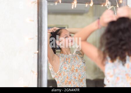 Ritratto di una ragazza adolescente con capelli neri crespi guardando lo specchio e facendo un ponytail. Foto Stock