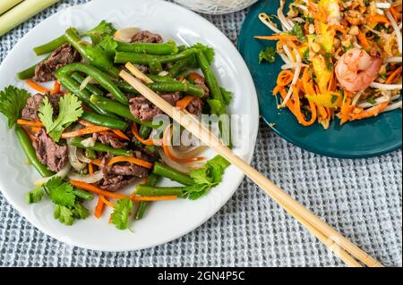 Due piatti sul tavolo con tradizionale pasto vietnamita e bacchette. Manzo fritto con fagiolo verde, carota e cipolla e insalata di mango con gamberi. Foto Stock