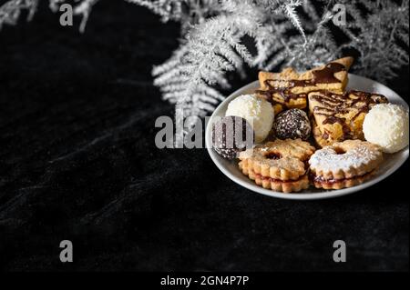 Mucchio di biscotti di natale su piatto bianco su sfondo di velluto nero con ramoscello bianco bracken. Foto Stock