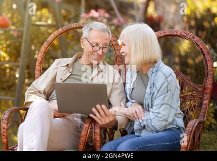 Coppia felice anziani rilassarsi in sedie di vimini all'aperto e utilizzando un computer portatile, navigare in internet o fare videochiamate online, spazio copia Foto Stock