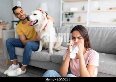 Signora caucasica malata che ha freddo, soffia il naso, starnutisce in carta tissue, il suo ragazzo arabo con cane da compagnia sullo sfondo Foto Stock