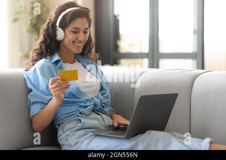 Shopping online. Felice signora caucasica con computer portatile e carta di credito seduta sul divano a casa, acquisto di merci in negozio web Foto Stock