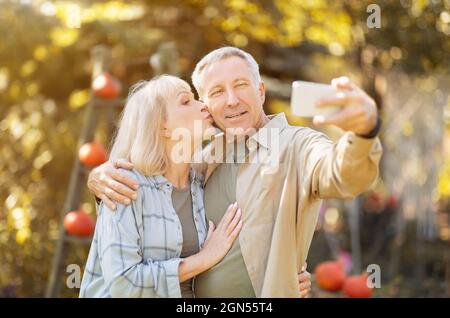 Coppia anziana che passa il tempo in giardino soleggiato insieme, prendendo selfie, donna baciando marito in guancia Foto Stock