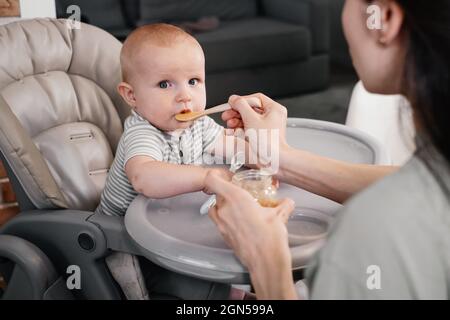 Prima pappa per bambini pura. Madre che dà il cucchiaio di verdure o frutta nella sedia di alimentazione a casa. Bambino carino che cerca di mangiare da solo. Sostegno sano Foto Stock