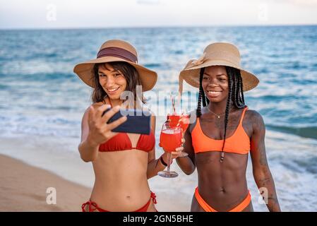 Giovane donna multirazziale sottile in bikini e cappelli clacking occhiali con bevande e prendere selfie sul telefono cellulare mentre godendo vacanze estive sulla spiaggia Foto Stock