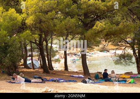 Kosirina, Murter, Croazia - 24 agosto 2021: Persone che riposano all'ombra dei pini sulla spiaggia e altri che nuotano in un mare Foto Stock