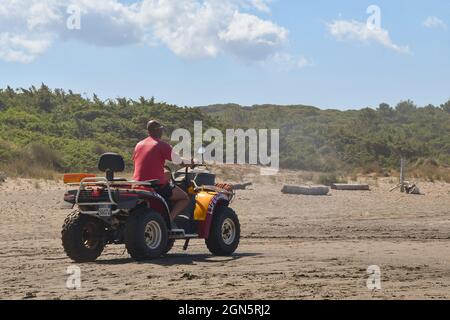 Un bagnino su un quad che pattina la spiaggia di sabbia sulla costa toscana con le dune coperte di vegetazione mediterranea in una giornata ventosa, Livorno Ev. Foto Stock