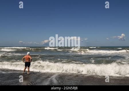 Vista posteriore di un uomo di mezza età in piedi sulla riva del mare mosso con grandi onde in estate, Marina di Castagneto Carducci, Livorno, Toscana, Italia Foto Stock