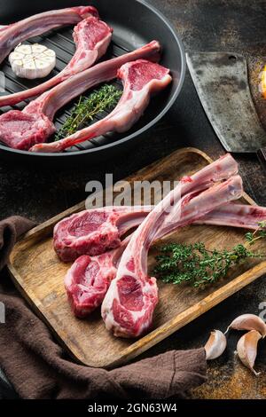 Set di braciole di agnello crudo, con ingredienti, su una padella in ghisa, su un vecchio sfondo rustico scuro Foto Stock