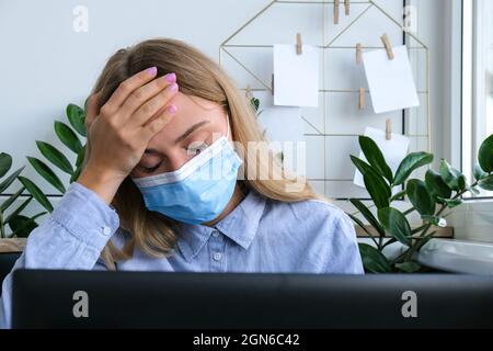 Stanco sonnolento giovane donna caucasica lavoratore indossare maschera medica al posto di lavoro scrivania. Lavoro eccessivo e privazione del sonno. Mal di testa Freelancer in depressione. Freddo e malato sul posto di lavoro Foto Stock