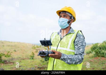 Operatore di drone con casco di sicurezza e maschera facciale che opera drone con telecomando - concetto di ingegnere che fa il rilevamento aereo utilizzando UAV durante Foto Stock