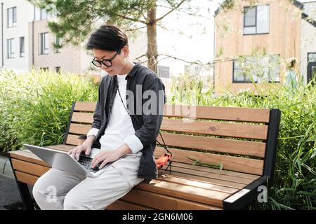 Ritratto orizzontale medio della giovane donna asiatica moderna che indossa abiti casual con occhiali seduti su panca all'aperto facendo un po 'di lavoro freelance utilizzando un computer portatile Foto Stock