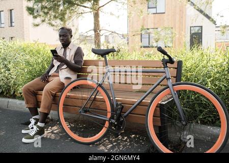 Lungo scatto orizzontale del giovane uomo afro-americano moderno che indossa un abito casual seduto rilassato su una panca di legno texting qualcosa sullo smartphone Foto Stock