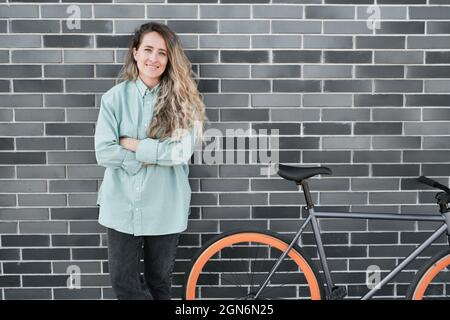 Attraente donna caucasica con lunghi capelli biondi in piedi con le braccia incrociate contro il muro grigio di mattoni all'aperto con la bicicletta parcheggiata accanto al suo sorridente a macchina fotografica Foto Stock