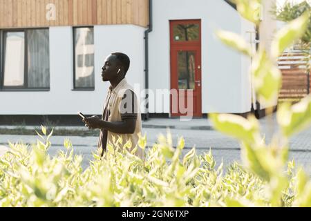 Scatto orizzontale laterale di un giovane afroamericano che cammina all'aperto con smartphone che ascolta musica utilizzando auricolari wireless Foto Stock