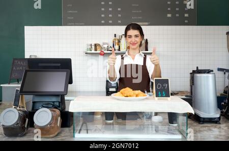 Il giovane negoziante caucasico solleva i pollici davanti al bancone di un bar. Atmosfera mattutina in una caffetteria. Foto Stock