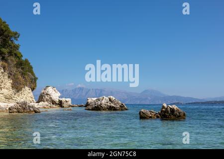 Scogli scenografici in acqua blu sulla spiaggia di ciottoli del Mar Ionio con montagne lontane e cielo luminoso. Natura dell'isola di Lefkada in Grecia. Vacanze estive tr Foto Stock