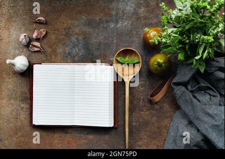 Vista dall'alto del libro di cucina bianco con grembiule, cucchiaio di legno e ingredienti freschi di aglio italiano, basilico e pomodori biologici.