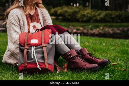 Ragazza in giacca a maglia, gonna borgogna e scarpe in pelle con zaino è seduto su erba verde nel parco dopo la scuola. Moda autunnale. Foto Stock