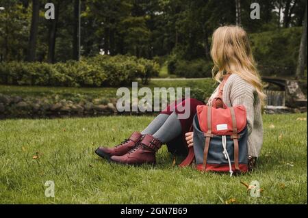 Ragazza in giacca a maglia, gonna borgogna e scarpe in pelle con zaino è seduto su erba verde nel parco dopo la scuola. Moda autunnale. Foto Stock